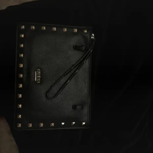 Svart plånbok med nitar från Guess - Snygg svart kuvertväska från Guess i skinn . Väskan har även ett handledsband!❣️