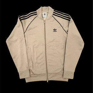 Beige Adidas ziptröja med svarta ränder - Säljer en beige Adidas ziptröja med klassiska svarta ränder längs ärmarna och broderad logga på bröstet. Tröjan har hel dragkedja, ribbade muddar och två fickor framtill. Perfekt för en sportig och stilren look.