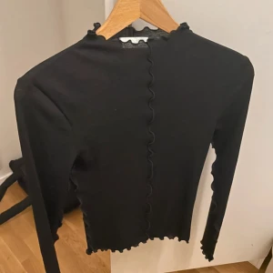 Svart långärmad topp från LeGer - Säljer en svart långärmad topp från LeGer med vågiga kanter längs halsringning, ärmslut och nederkant. Toppen är ribbad och har en slim passform som sitter snyggt på kroppen. Perfekt för dig som gillar stilrena och trendiga plagg.