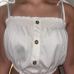 Vit linnetopp med knappar från H&M - Supersöt vit linnetopp från H&M med smock upptill och nertill, knytbara axelband och tre dekorativa knappar framtill. Luftig och perfekt för varma dagar, tillverkad i mjukt bomullstyg. Enkel att styla med jeans eller kjol.