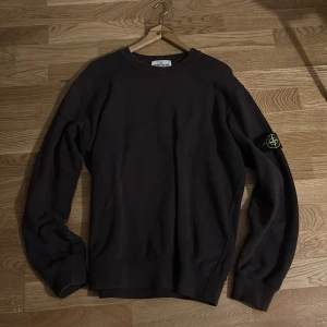 Mörkbrun sweatshirt från Stone Island - Snygg mörkbrun sweatshirt från Stone Island med klassisk rund hals och ikonisk patch på vänster ärm. Tröjan har ribbade muddar och är tillverkad i mjuk bomull. Perfekt för dig som gillar streetwear och vill ha något stilrent med cool detalj.