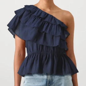 volang topp - Snygg marinblå topp med volanger och oneshoulder-design. Toppen har flera lager volanger över axeln och en smickrande, lätt utsvängd midja. Perfekt för dig som vill sticka ut med en cool och trendig look.