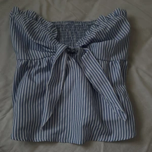 Blåvit randig linnetopp med volang - Supersöt blåvit randig linnetopp med volangkant upptill och smockad rygg. Toppen har knyt framtill och är ärmlös, perfekt för varma dagar. Materialet känns lätt och luftigt, och den har en feminin och fräsch vibe.