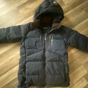 Mörkblå pufferjacka från Sail Racing - Snygg mörkblå pufferjacka från Sail Racing med huva och två bröstfickor med dragkedja. Jackan har quiltad design, hög krage och logga på bröstet. Perfekt för kalla dagar och riktigt skön att bära.                Storlek: 160