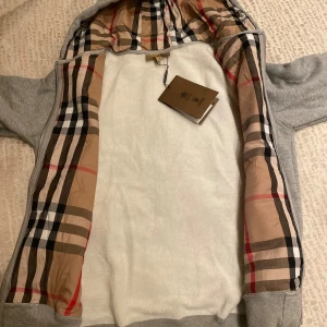 Grå hoodie från Burberry med rutig insida - Snygg grå hoodie från Burberry i storlek M. Insidan av luvan och öppningen har det klassiska Burberry-rutiga mönstret i beige, svart, vitt och rött. Tröjan har dragkedja framtill och ribbade muddar. Perfekt för dig som gillar exklusiva streetwear-vibbar.