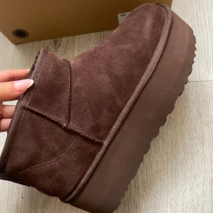 Bruna UGG boots med platåsula - Säljer ett par bruna UGG boots i mocka med tjock platåsula. Insidan är fodrad med mjuk ull för extra värme. Perfekta för kalla dagar och ger en snygg look. Klassisk UGG-logga bak på hälen 