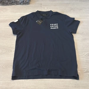 Marinblå Peak Performance piké XXL - Snygg marinblå pikétröja från Peak Performance i storlek XXL. Tröjan har klassisk krage, korta ärmar och vita tryck på bröstet och ärmen. Tillverkad i mjuk bomull med knappslå framtill. Perfekt för dig som gillar stilren design och sportig vibe.