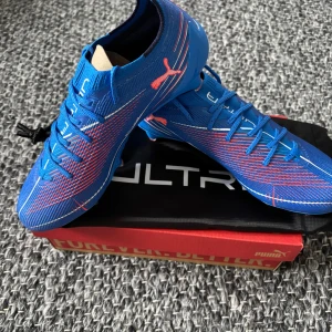 Blåa Puma Ultra fotbollsskor - Säljer ett par grymma blåa Puma Ultra fotbollsskor med snygga röda och vita detaljer. Skorna har snörning, låg profil och mönstrad ovandel i syntetmaterial för bästa grepp och känsla på planen. Sulan har dobbar för optimalt fäste på gräs. Helt oanvända! Nypris 2700