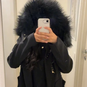 Svart parkajacka från Dennis Maglic - Svart parkajacka från Dennis Maglic med stor huva kantad av fluffig svart fuskpäls. Jackan har snygga gulddetaljer, dragkedja framtill och dekorativa spännen. Perfekt inför höst/vinter. Köpt för 1799kr och säljs inte längre i hemsidan. 