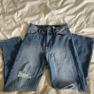 Baggy blå jeans från &Denim, strl 34 - Baggy jeans i klassisk blå denim från &Denim, storlek 34. Modellen har låg midja och raka ben med slitna detaljer vid knäna. Jeansen har fem fickor, knapp och dragkedja framtill. Perfekt för dig som gillar avslappnad stil.