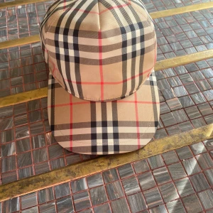 Burberry rutig keps med logga - Snygg keps från Burberry med klassiskt rutigt mönster i beige, svart, vitt och röda linjer. Justerbar baktill med svart plastspänne och tydlig Burberry London England-logga. Perfekt accessoar för dig som gillar street och designer.