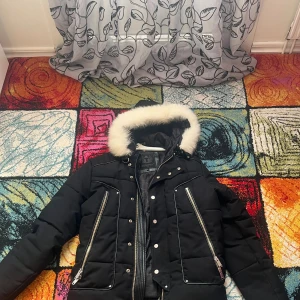 Svart pufferjacka med fuskpäls från Horspist - Svart pufferjacka från Horspist med vit fuskpäls runt huvan, långa ärmar och flera silvriga dragkedjor framtill. Jackan har quiltad design, tryckknappar och en snygg urban look. Perfekt för kalla dagar med sin tjocka vaddering och coola detaljer. Bara dragkedjan funkar inte.OBS KVITTO FINNS INTE