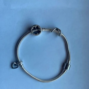 Pandora silverarmband med berlocker - Säljer ett snyggt Pandora armband i silver med tre berlocker: en fyrklöver, ett oändlighetstecken och ett hjärta. Armbandet har klassisk design och berlockerna ger en personlig touch. Perfekt för dig som gillar att mixa och matcha smycken. Längden är 18cm 