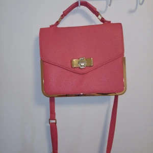 Rosa axelväska med gulddetaljer - LÄS HÄR!! Snygg rosa axelväska med guldfärgade metalldetaljer och ett elegant spänne framtill. Väskan har både handtag och justerbar axelrem, samt ett innerfack med dragkedja. Perfekt för dig som vill ha en stilren och trendig accessoar. Men som ni se på sista bilden så har den lite fläder inuti väskan och nån fläk bak på väskan. Såå snygg ❤️ skriv för fler frågor 