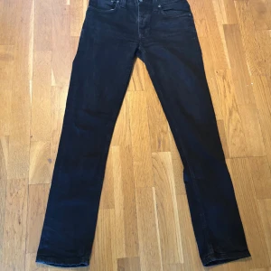Svarta jeans från Nudie Jeans, W32  - Snygga svarta jeans från Nudie Jeans med klassisk femficksdesign och diskret broderi på bakfickorna. Jeansen har rak passform och är tillverkade i mjukt bomullstyg. Perfekta för dig som gillar stilrena och tidlösa jeans. 
