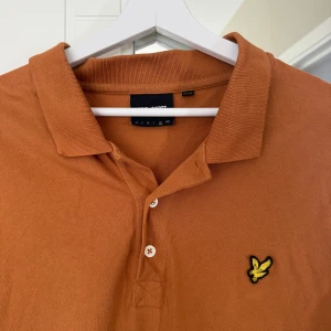 Brun pikétröja från Lyle & Scott - Säljer en snygg brun pikétröja från Lyle & Scott med klassisk krage, korta ärmar och två knappar framtill. Tröjan har den ikoniska gula fågel-loggan broderad på bröstet och är gjord i mjuk bomull. Perfekt för en stilren och avslappnad look.