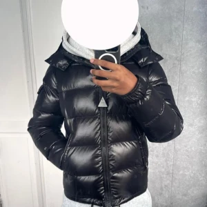 Svart Moncler maya, sitter som M / L - Säljer en svart pufferjacka från Moncler med glansig finish och huva. Jackan har Moncler-logga på ärmen, praktiska fickor och dragkedja framtill. Perfekt för dig som vill ha en snygg och varm jacka med street vibe under kalla dagar.              OBS = hjälper en polare