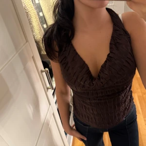 Brun omlott halterneck topp - Säljer en brun omlott halterneck topp med snygg v-ringning och öppen rygg. Toppen har ett strukturerat tyg med veckad effekt som ger cool textur. Perfekt att styla med jeans eller kjol för en trendig look.