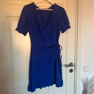 Blå omlottklänning med volang - Supersnygg blå omlottklänning med kort ärm och volangkant nertill. Klänningen har knyt i midjan och en lätt strukturerad yta. Perfekt för dig som gillar en feminin och trendig look med v-ringning och puffärm.