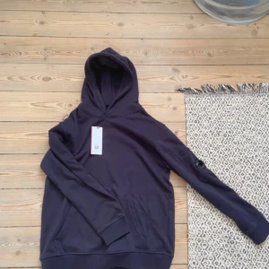 Mörkblå hoodie från C.P. Company - Säljer en mörkblå hoodie från C.P. Company med klassisk huva och känguruficka. Ärmen har den ikoniska linsen med logga. Tröjan är i mjuk bomull och har långa ärmar, perfekt för en chill och stilren look.