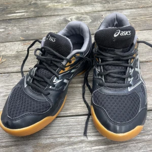 Svarta Asics Upcourt 4 sneakers - Säljer ett par Asics Upcourt 4 sneakers i svart med grå och silverdetaljer samt gummifärgad sula. Skorna har snörning och meshpartier för extra andningsförmåga. Perfekta för inomhussport eller träning. Snygg och sportig design med bekväm passform.