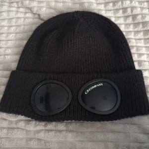 Svart C.P. Company goggle-mössa - Svart ribbstickad mössa från C.P. Company med ikoniska goggle-linser framtill på uppviket. Tillverkad i mjuk ull och har en klassisk, tight passform. Perfekt accessoar för att sticka ut i vinterkylan.