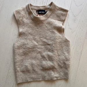 Beige stickad väst från Pieces - Supersnygg beige stickad väst från Pieces med ribbad struktur och rund hals. Modellen är croppad och har breda axelpartier utan ärmar, vilket ger en modern och clean look. Perfekt att styla över en skjorta eller t-shirt för en trendig vibe.