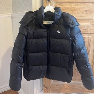 Svart pufferjacka från Calvin Klein - Säljer en svart pufferjacka från Calvin. Inte mycket använd och finns inga deftekter, hål, repor eller liknande. Väldigt varm och har luva som går att ta av om man vill. Storlek S.