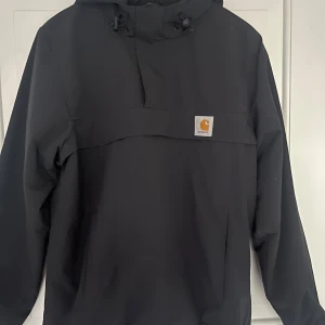 Fodrad vindjacka från Carhartt WIP - Svart vindjacka med fodring inuti från Carhartt WIP med huva. Jackan har en klassisk logopatch på bröstet och är tillverkad i slitstark polyester. Perfekt för dig som gillar streetwear och enkel stil. Sitter lite mer som M. Perfekt till höst och vinter. Hör av dig vid funderingar 👍
