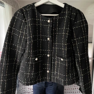 Svart rutig kavajjacka - Svart kavajjacka i rutigt mönster med vita linjer. Jackan har pärlknappar framtill och två fickor. Går att använda som både jacka och kofta, den är något kortare i modellen. 
