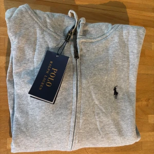 Grå hoodie från Polo Ralph Lauren - Snygg ljusgrå hoodie från Polo Ralph Lauren med dragkedja, huva och känguruficka. Klassisk broderad logga på bröstet och ribbade muddar. Tillverkad i mjuk bomullsmix, perfekt för chill dagar eller när du vill ha en clean look.