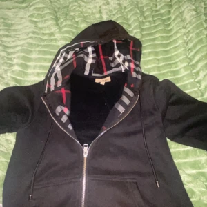 Svart Burberry hoodie med dragkedja - Svart hoodie från Burberry med klassiskt rutigt foder i huvan i svart, vitt och rött. Tröjan har hel dragkedja framtill, snörning i huvan och två fickor. Perfekt för dig som vill ha en stilren och lyxig streetwear-look.