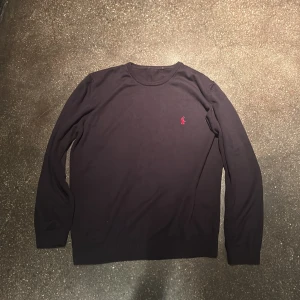 Svart tröja från Ralph Lauren - Snygg svart  långärmad tröja från Ralph Lauren med klassisk rund hals och röd broderad logga på bröstet. Tröjan är i mjuk bomull och har ribbade muddar vid ärmslut och nederkant. Perfekt för en stilren och avslappnad look.