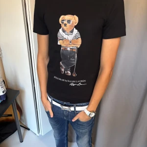 Svart Ralph Lauren T-shirt  - Svart t-shirt från Polo Ralph Lauren med ikoniskt Polo Bear-tryck framtill. Helt ny med tags. Klassisk passform, rund hals och korta ärmar. Tillverkad i mjuk bomull som känns skön mot huden. Perfekt statement-plagg för dig som gillar streetstyle och snygga prints. Hör gärna av er vid frågor!🙌🙌