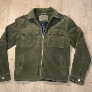 Olivgrön Human Scales mockajacka S - Olivgrön mockajacka från Human Scales, modell Rick storlek S. Fodret är mörkblått och jackan har klassisk krage och knappdetaljer vid ärmslut. Perfekt för dig som gillar stilren och cool look. Använd i nyskick. Meddela för frågor!