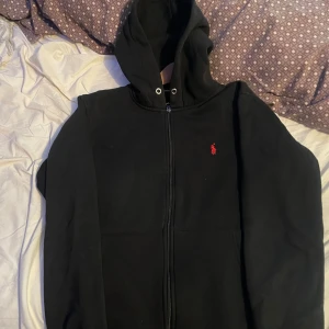 Svart hoodie från Ralph Lauren - Svart hoodie från Ralph Lauren med dragkedja och huva. Klassisk röd broderad logga på bröstet. Mjuk och skön bomullskvalitet, perfekt för chill dagar. Snygg och enkel design med fickor framtill.