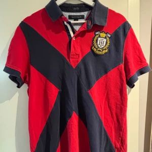 Röd och marinblå Tommy Hilfiger piké - Snygg röd och marinblå t-shirt från Tommy Hilfiger med broderad crest på bröstet och stora siffran 8 på ryggen. T-shirten har korta ärmar, krage och är gjord i mjuk bomull. Perfekt för dig som gillar sportig och klassisk stil.