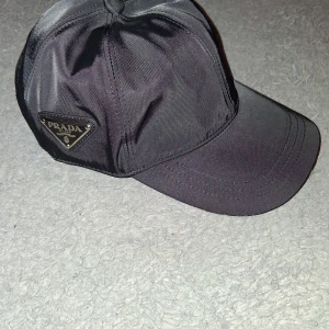 Svart keps från Prada i nylon - Snygg svart keps från Prada i glansigt nylonmaterial med justerbar rem bak. Klassisk böjd skärm och ikonisk metallogo på sidan. Perfekt accessoar för en clean och lyxig streetstyle-look.