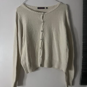 Beige kofta från Indiska med knappar - Snygg beige kofta från Indiska i storlek M. Den är stickad i mjukt material och har långa ärmar samt pärlemorskimrande knappar framtill. Perfekt för lager-på-lager och har en klassisk rund halsringning och ribbade muddar.