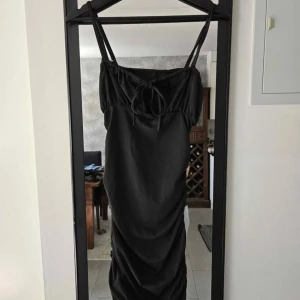 Svart långklänning med snörning - Svart långklänning med smala axelband och snygg snörning framtill vid bysten. Klänningen har en åtsittande passform och rynkade detaljer längs sidorna som ger en cool siluett. Perfekt för dig som gillar stilrena och trendiga plagg.