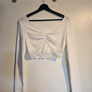 Vit croppad långärmad topp med spets - Säljer en vit croppad topp med långa ärmar och ribbat tyg. Toppen har v-ringning, rynkdetalj framtill och en snygg spetskant nedtill. Perfekt för dig som gillar romantiska detaljer och en fräsch look.