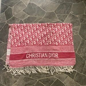 Röd halsduk från Christian Dior - Snygg och stor halsduk från Christian Dior i röd och vitt med logotypmönster och tydlig text. Halsduken har fransar i ändarna och ett klassiskt, rektangulärt snitt. Perfekt för dig som vill sticka ut med ett lyxigt statement-plagg.
