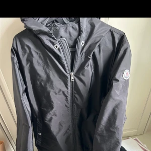 Svart vindjacka från Moncler - Svart vindjacka från Moncler med huva och dragkedja framtill. Jackan har Moncler-logga på ärmen och praktiska fickor med knappstängning. Perfekt för blåsiga dagar och enkel att matcha med allt.