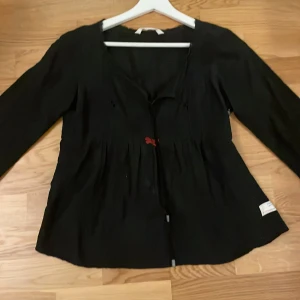 Svart blus med rosett från Odd Molly - Supersöt svart blus från Odd Molly med lång ärm och en liten röd rosett framtill. Köpt på loppis och har aldrig använt den själv 