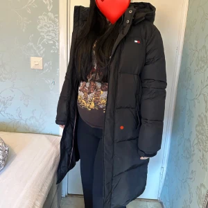 Svart lång pufferjacka Tommy Hilfiger - Säljer en svart lång pufferjacka från Tommy Hilfiger med huva och diskret logga på bröstet. Jackan har dragkedja framtill och är riktigt fluffig och varm, perfekt för kalla dagar. Passformen är normal och jackan har långa ärmar.