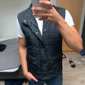 Svart quiltad dunväst från Bläck - Snygg svart quiltad dunväst med knappar och dragkedja framtill. Västen har ståkrage, två fickor med lock och en bröstficka med dragkedja. Perfekt för lager-på-lager och ger en cool look till jeans och t-shirt.