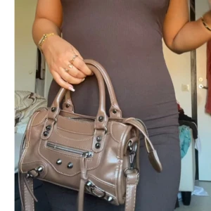 Brun axelväska i skinnstil - Cool brun axelväska med både handtag och lång axelrem. Väskan har snygga metalldetaljer, dragkedja framtill och dekorativa spännen. Perfekt storlek för det viktigaste och ger en edgy vibe till din outfit.