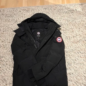 Svart dunjacka från Canada Goose - Säljer en svart dunjacka från Canada Goose med huva och klassisk logga på ärmen. Jackan har dragkedja och knappar framtill, samt en stilren, vadderad design som håller dig varm under kalla dagar. Perfekt för dig som vill ha en snygg och funktionell vinterjacka.