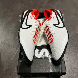 Nike vita fotbollsskor med röd detalj - Säljer ett par grymma vita Nike fotbollsskor med svarta och röda detaljer. Skorna har snörning, rund tå och platt sula med dobbar för bästa grepp på planen. Ovandelen är i syntetmaterial och insidan är fodrad i rött för extra stilpoäng. Pris kan diskuteras!