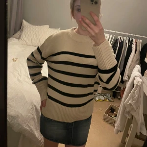 Randig beige stickad tröja - Säljer en beige stickad tröja med svarta horisontella ränder. Tröjan har rund halsringning, breda ribbade muddar och lång ärm. Oversized och mysig passform, perfekt för lager-på-lager under kyliga dagar.